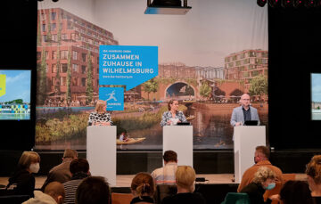 © IBA Hamburg / Daniela Hoffmann Drei Redner:innen stehen an Pulten vor Publikum auf einer Bühne