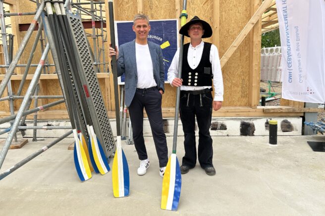 © IBA Hamburg / Anke Hansing Kay Gätgens und ein Zimmermann in traditioneller Kleidung mit Rudern in den Händen posieren vor einer Wand aus Holzelementen