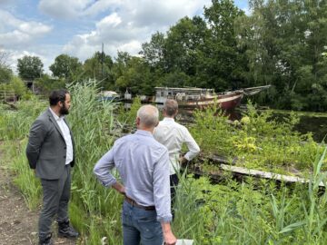 © IBA Hamburg / Arne von Maydell Drei Männer stehen im Schilf am Ufer eines Kanals und blicken auf ein altes, verwittertes Boot mit der Aufschrift Isabella