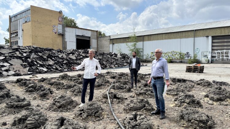© IBA Hamburg / Arne von Maydell Drei Männer stehen auf aufgewühltem, dunklem Erdboden vor einem Schutthaufen und alten Industriegebäuden mit Graffiti