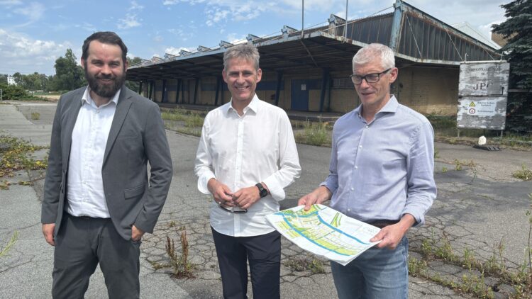 © IBA Hamburg / Arne von Maydell Drei Männer stehen lächelnd vor einer verlassenen, offenen Lagerhalle, einer hält einen Lageplan in der Hand