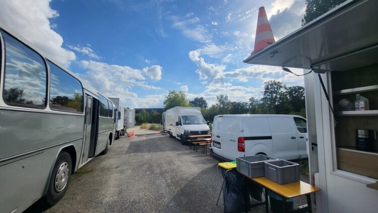 © IBA Hamburg / Sebastian Brose Ein Bus und mehrere weiße Transportfahrzeuge stehen auf dem Gelände. Rechts ist ein Versorgungswagen mit geöffneter Ausgabeklappe zu sehen.