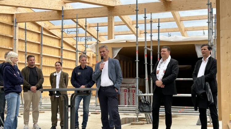 © IBA Hamburg / Anke Hansing Eine Person spricht vor Gästen im Rohbau des neuen Vereinshauses, umgeben von sichtbarer Holzkonstruktion und Baugerüsten.