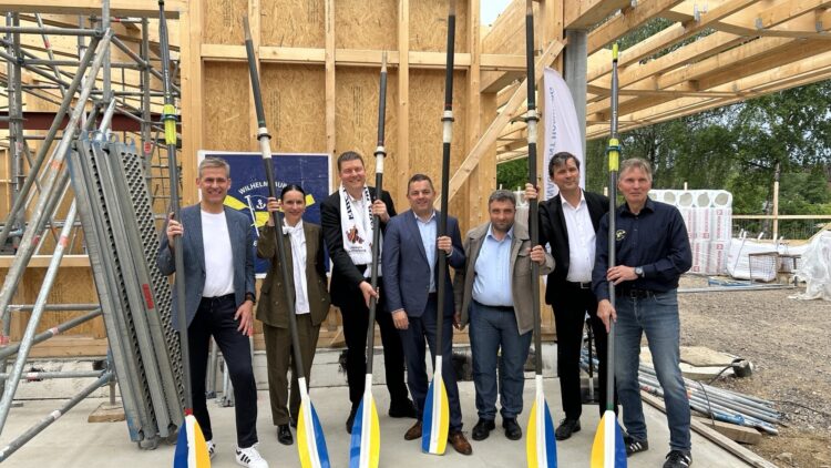 © IBA Hamburg / Anke Hansing Mehrere Personen posieren mit Ruderriemen vor der Holzkonstruktion des neuen Vereinshauses auf der Baustelle.