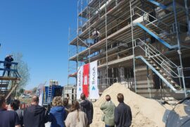 © IBA Hamburg / Arne von Maydell Menschen auf einer SAGA-Baustelle, auf einem Gerüst hält ein Zimmermann eine Rede