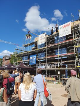 © IBA Hamburg Menschen schauen auf einen Rohbau, oben sind die Richtfestkrone und zwei Handwerker zu sehen