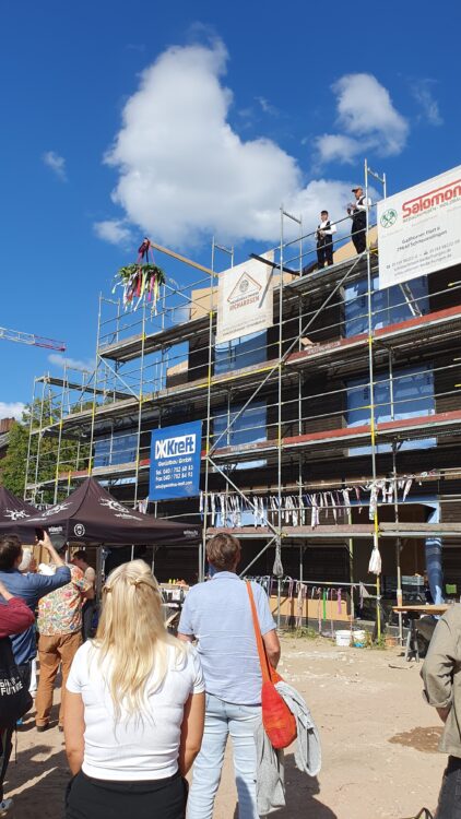 © IBA Hamburg enschen schauen auf einen Rohbau, oben sind die Richtfestkrone und zwei Handwerker zu sehen