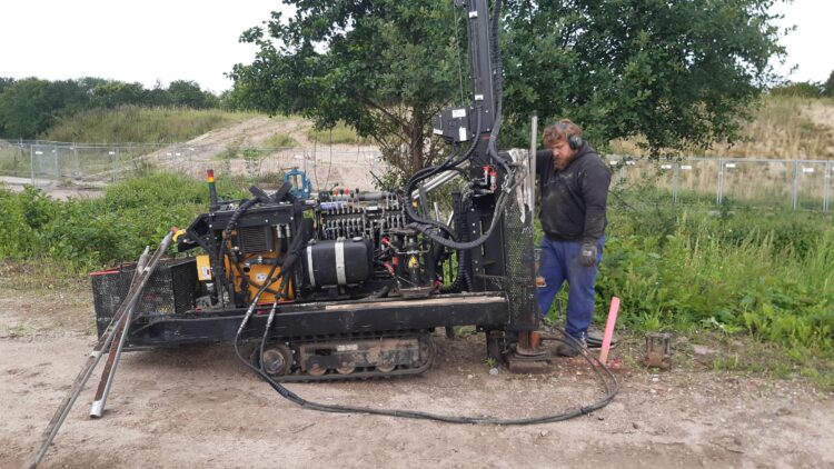© IBA Hamburg / Thomas Waldau Person in Arbeitskleidung neben Bohrgerät zur Bodenuntersuchung; umgeben von Werkzeugen, im Freien.