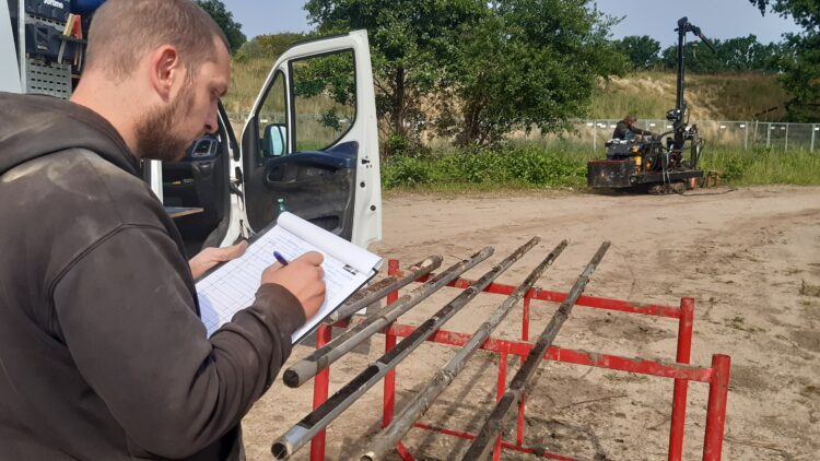 © IBA Hamburg / Thomas Waldau Person schreibt auf Klemmbrett neben Bohrkernhalterung und offenem weißen Transporter; im Hintergrund Grün und Maschinen.