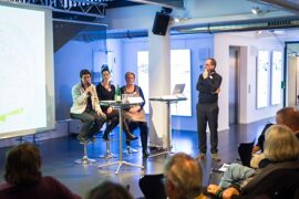 Vier Personen sitzen mit Mikrofon vor einer Leinwand vor Publikum