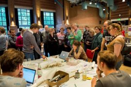 Menschen stehen um einen Tisch, auf dem Karten, Zeichnungen und Notizen liegen